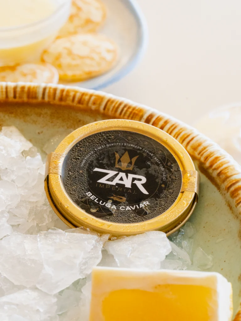 Lata dorada de caviar Beluga ZAR Imperial sobre hielo picado en un cuenco de cerámica, con blinis y mantequilla desenfocados al fondo