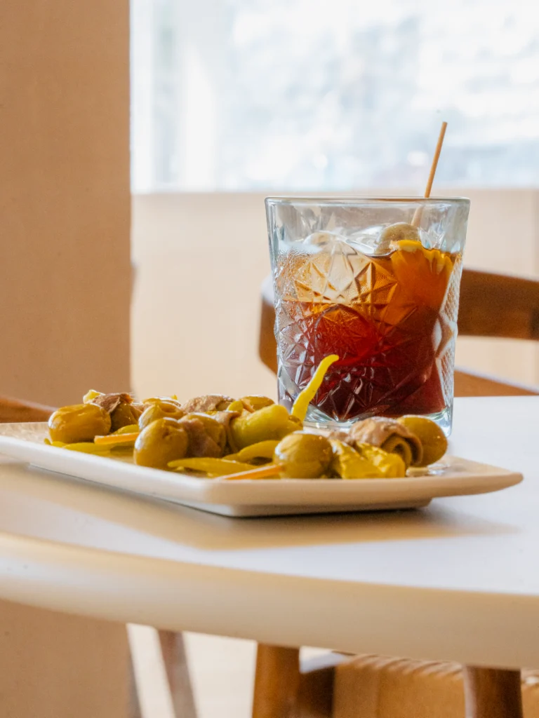 Vista a ras de mesa de un plato de gildas con anchoa y guindilla y un vaso de vermut con hielo y naranja ante una ventana luminosa