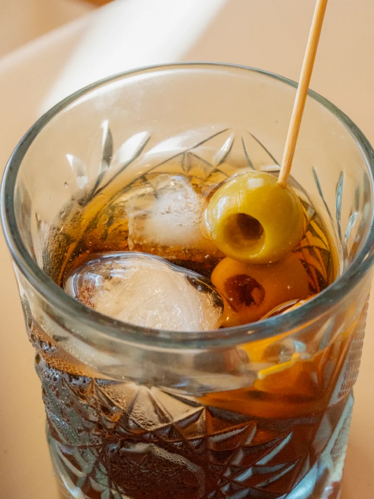 Primer plano de un vaso de cristal tallado con vermut, hielo, una aceituna con palillo de madera y piel de naranja